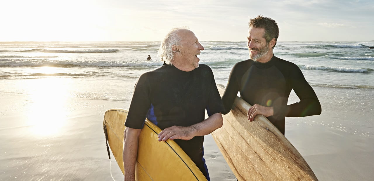 A father and son laugh together while carrying surfboards away from the beach.