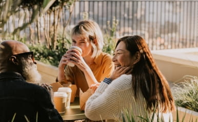 Three colleagues sit outside on a sunny day and enjoy a takeaway lunch and some hot drinks. They are comfortable with each other, having a light hearted discussion.